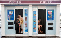 Young girls joke with a boy inside his malfunctioning photo booth.